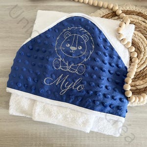 Peut inclure: Serviette à capuche bleue avec une bordure blanche et un lion brodé argenté avec le nom Milo.