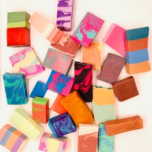 May include: A collection of handmade soap bars in various colors and patterns. The soaps feature layered designs, swirls, and marbled effects in shades of pink, blue, green, orange, and brown. Some bars have a rectangular shape, while others are more square.