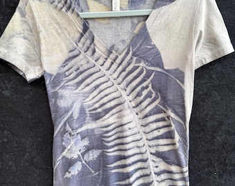 May include: A short-sleeved, V-neck t-shirt with a botanical print. The design features a fern and leaf pattern in shades of blue and off-white. The shirt is hanging on a light green hanger.