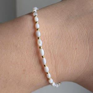 Könnte beinhalten: Ein zartes Armband mit kleinen, oval geformten weißen Perlen, die mit goldfarbenen Abstandshaltern versehen sind. Das Armband ist auf einem flexiblen Band aufgereiht und für das Handgelenk konzipiert. Die Perlen haben einen dezenten Glanz.