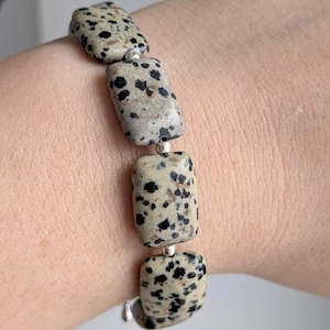 Könnte beinhalten: Ein Armband mit rechteckigen, gesprenkelten Steinen in Beige und Schwarz. Die Steine sind mit silberfarbenen Verbindern verbunden. Das Armband wird am Handgelenk getragen und zeigt das Design und die Farbe des Schmucks.