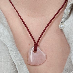 Könnte beinhalten: Halskette mit herzförmigem Rosenquarz-Anhänger und burgunderfarbenem Band. Der Anhänger hat eine glatte, polierte Oberfläche und ein kleines Loch oben. Die Halskette wird über einem hellen Hemd getragen.