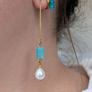 Könnte beinhalten: Ein goldener Ketten-Ohrhänger mit einer zylindrischen türkisfarbenen Perle und einem weißen Perlenanhänger. Am Ohr befinden sich zudem ein goldener Creolen-Ohrring und ein goldener Stecker mit zwei kleinen türkisfarbenen Perlen.