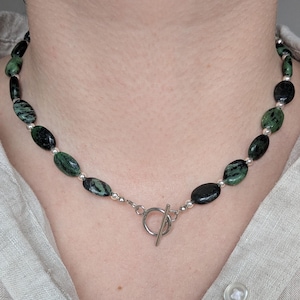 Könnte beinhalten: Halskette mit ovalen, dunkelgrünen und schwarzen Steinen, aufgereiht mit kleinen silbernen Perlen. Sie hat einen silbernen Knebelverschluss. Der Schmuck wird vor einem neutralen Hintergrund gezeigt.