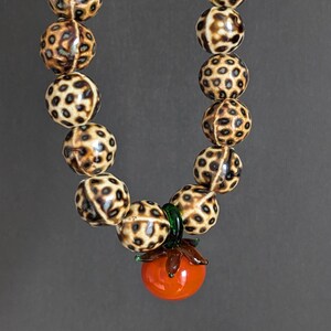 May include: A beaded necklace featuring large, round beads with a spotted pattern in shades of brown and cream. The necklace has a pendant of a red glass tomato with green leaves.