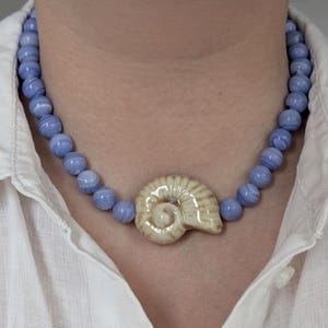 Könnte beinhalten: Eine Perlenkette mit runden, hellblauen, gebänderten Achatperlen und einem zentralen, cremefarbenen Keramikanhänger in Form eines Ammoniten. Die Kette wird um den Hals getragen und liegt auf einem weißen Hemd mit Kragen und sichtbarem Knopf.