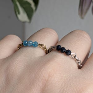 Könnte beinhalten: Zwei Kettenringe mit kleinen facettierten Perlen. Ein Ring besteht aus goldfarbenem Metall mit drei hellblauen Perlen, der andere aus silberfarbenem Metall mit drei dunkelblauen Perlen, beide an nebeneinander liegenden Fingern getragen.