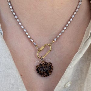 Könnte beinhalten: Eine Halskette mit einer Reihe kleiner, hellvioletter, ovaler Perlen. Die Kette hat in der Mitte einen goldfarbenen Karabinerverschluss aus Metall, an dem ein dunkelbrauner, durchscheinender Muschelanhänger aus Glas mit gerippter Oberfläche hängt.