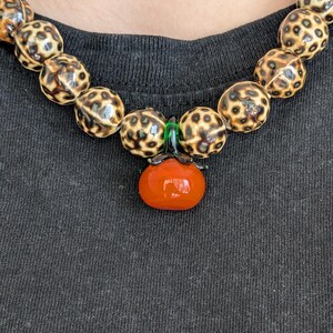 May include: A beaded necklace featuring large, round beads with a spotted pattern in shades of brown and cream. A pendant shaped like a small, orange tomato with green accents hangs from the necklace. The necklace is worn against a dark gray background.