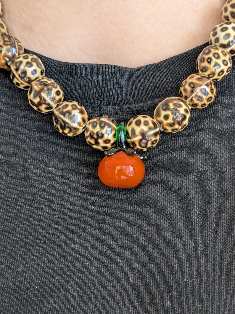 May include: A beaded necklace featuring large, round beads with a spotted pattern in shades of brown and cream. A pendant shaped like a small, orange tomato with green accents hangs from the necklace. The necklace is worn against a dark gray background.