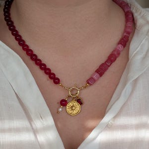 Könnte beinhalten: Eine zweifarbige Perlenkette mit goldenem Anhänger. Die Kette wechselt von dunkelroten zu rosa Perlen. Der goldene Anhänger hat ein Sternmuster, einen roten Edelstein und eine kleine Perle. Die Kette wird über einem weißen Hemd getragen.