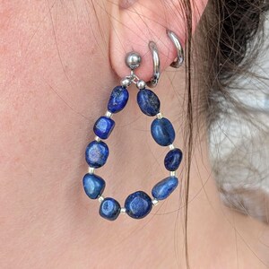 Könnte beinhalten: Ein tropfenförmiger Ohrring mit polierten, tiefblauen Lapislazuli-Steinen, die mit kleinen silberfarbenen Perlen durchsetzt sind. Der Ohrring ist an einem einfachen silberfarbenen Stecker befestigt, daneben sind zwei schlichte silberne Creolen.