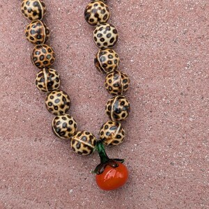 May include: A beaded necklace featuring round, spotted beads in shades of brown and tan. The necklace has a small, orange tomato pendant with green leaves. The beads are approximately 0.75 inches in diameter.