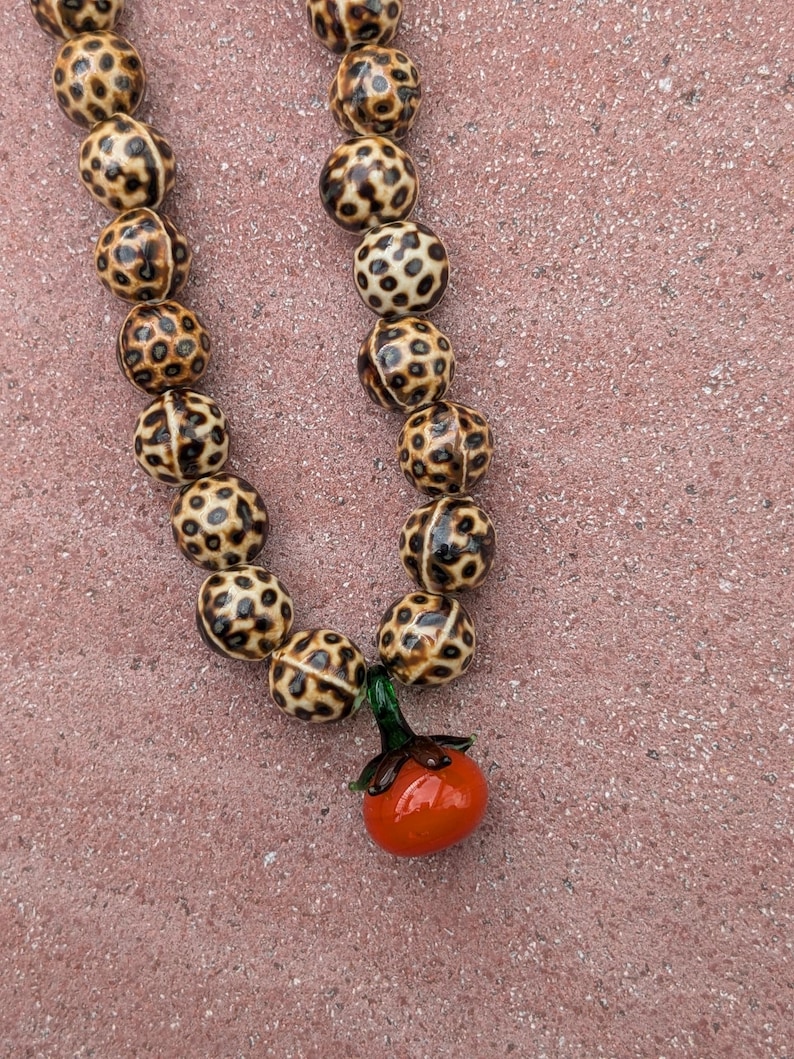 May include: A beaded necklace featuring round, spotted beads in shades of brown and tan. The necklace has a small, orange tomato pendant with green leaves. The beads are approximately 0.75 inches in diameter.