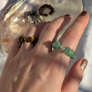 Könnte beinhalten: Eine Hand zeigt drei Edelsteinringe: einen mit Tigeraugensteinen, einen mit grünen Steinen und einen dritten mit dunkelroten Steinen. Ein vierter Ring mit klaren Steinen ist ebenfalls sichtbar. Die Ringe befinden sich auf einer weißen Oberfläche mit einer Muschel im Hintergrund.