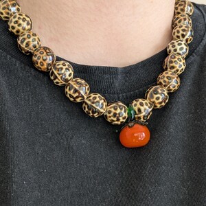 May include: A beaded necklace featuring large, round beads with a spotted pattern in shades of tan and brown. The necklace has a small, orange tomato-shaped pendant with a green accent. The necklace is worn against a black background.