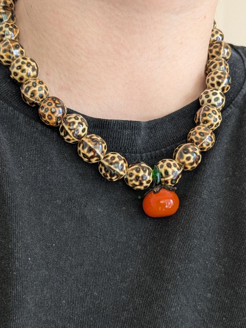 May include: A beaded necklace featuring large, round beads with a spotted pattern in shades of tan and brown. The necklace has a small, orange tomato-shaped pendant with a green accent. The necklace is worn against a black background.