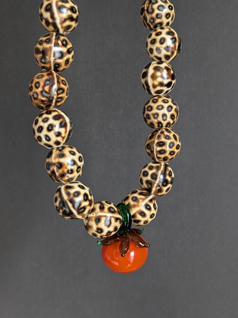 May include: A beaded necklace featuring large, round beads with a spotted pattern in shades of brown and cream. The necklace has a pendant of a red glass tomato with green leaves.