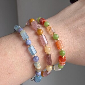 Könnte beinhalten: Drei Armbänder mit Perlen in Blau-, Lila-, Rosa-, Orange-, Grün- und Gelbtönen. Die Armbänder weisen eine Vielzahl von Perlenformen auf, darunter runde und rechteckige, mit goldfarbenen Abstandshaltern. Die Armbänder werden am Handgelenk getragen.