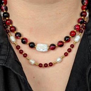 Könnte beinhalten: Zweisträngige Halskette mit rot-schwarz gesprenkelten Perlen, schwarzen Perlen und weißen Perlen. Der obere Strang hat eine große, rechteckige weiße Perle. Die Halskette ist auf goldfarbenem Draht aufgefädelt.