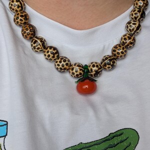 May include: A beaded necklace featuring large, round beads with a spotted pattern in shades of brown and cream. A small, red tomato pendant with green accents hangs from the necklace. The necklace is worn over a white t-shirt.