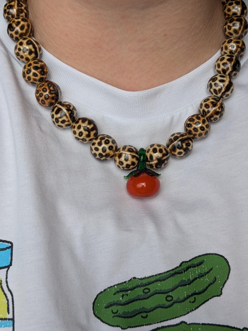 May include: A beaded necklace featuring large, round beads with a spotted pattern in shades of brown and cream. A small, red tomato pendant with green accents hangs from the necklace. The necklace is worn over a white t-shirt.