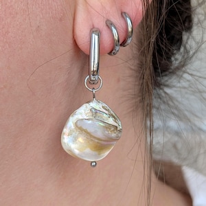 Könnte beinhalten: Ein silberfarbener Metall-Creolen-Ohrring mit einem baumelnden, unregelmäßigen, irisierenden Muschel-Anhänger in Weiß-, Creme- und Goldtönen. Der Ohrring wird zusammen mit zwei kleineren, schlichten silberfarbenen Creolen getragen.