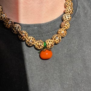May include: A beaded necklace featuring large, round beads with a spotted pattern in shades of brown and cream. The necklace has an orange pendant shaped like a tomato. The necklace is worn against a gray background.