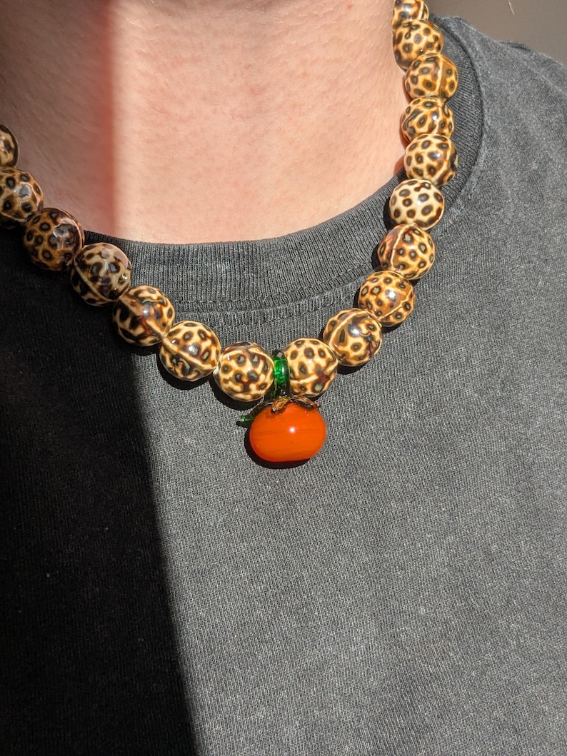 May include: A beaded necklace featuring large, round beads with a spotted pattern in shades of brown and cream. The necklace has an orange pendant shaped like a tomato. The necklace is worn against a gray background.