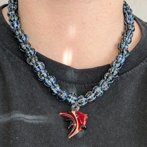 Könnte beinhalten: Eine Halskette mit Anhänger. Die Halskette hat dunkelblaue und schwarze Glasperlen. Der Anhänger ist ein roter, schwarzer und klarer Glasfisch mit einem silberfarbenen Bügel.