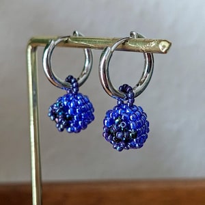 Könnte beinhalten: Ein Paar Creolen mit einem Cluster aus blauen Perlen. Die Ohrringe haben silberfarbene Creolen und einen kleinen, runden Cluster aus blauen Perlen, der an jeder Creole hängt. Die Perlen haben ein glänzendes, irisierendes Finish.