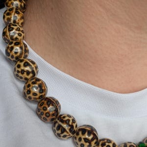 May include: A beaded necklace featuring large, round beads with a spotted pattern in shades of brown and cream. The necklace is worn against a white shirt, highlighting the contrasting colors and textures of the jewelry.