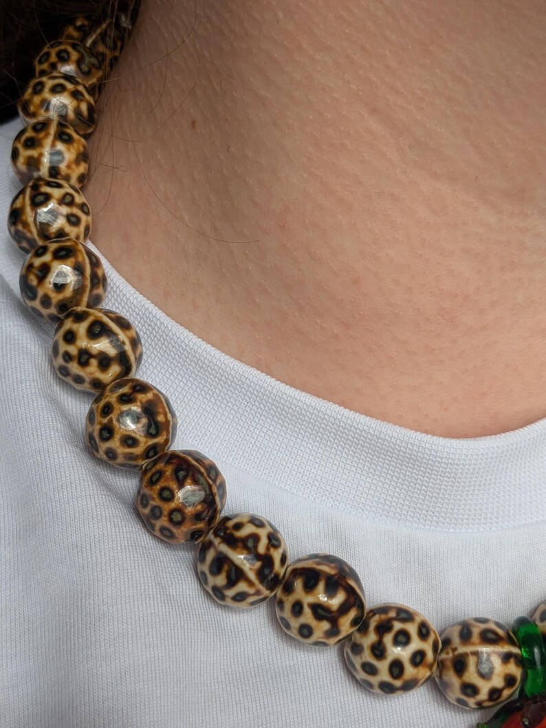 May include: A beaded necklace featuring large, round beads with a spotted pattern in shades of brown and cream. The necklace is worn against a white shirt, highlighting the contrasting colors and textures of the jewelry.