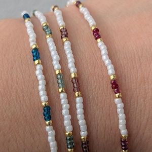 Könnte beinhalten: Vier Armbänder mit Perlen aus weißen, blauen, grünen, lila und goldenen Perlen. Die Armbänder sind auf einer weißen Schnur aufgefädelt. Die Armbänder liegen eng beieinander, und die Perlen sind klein und gleichmäßig groß.