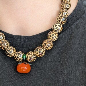 May include: A beaded necklace featuring large, round beads with a spotted pattern in shades of brown and cream. The necklace has a pendant of a small, orange tomato with green accents.