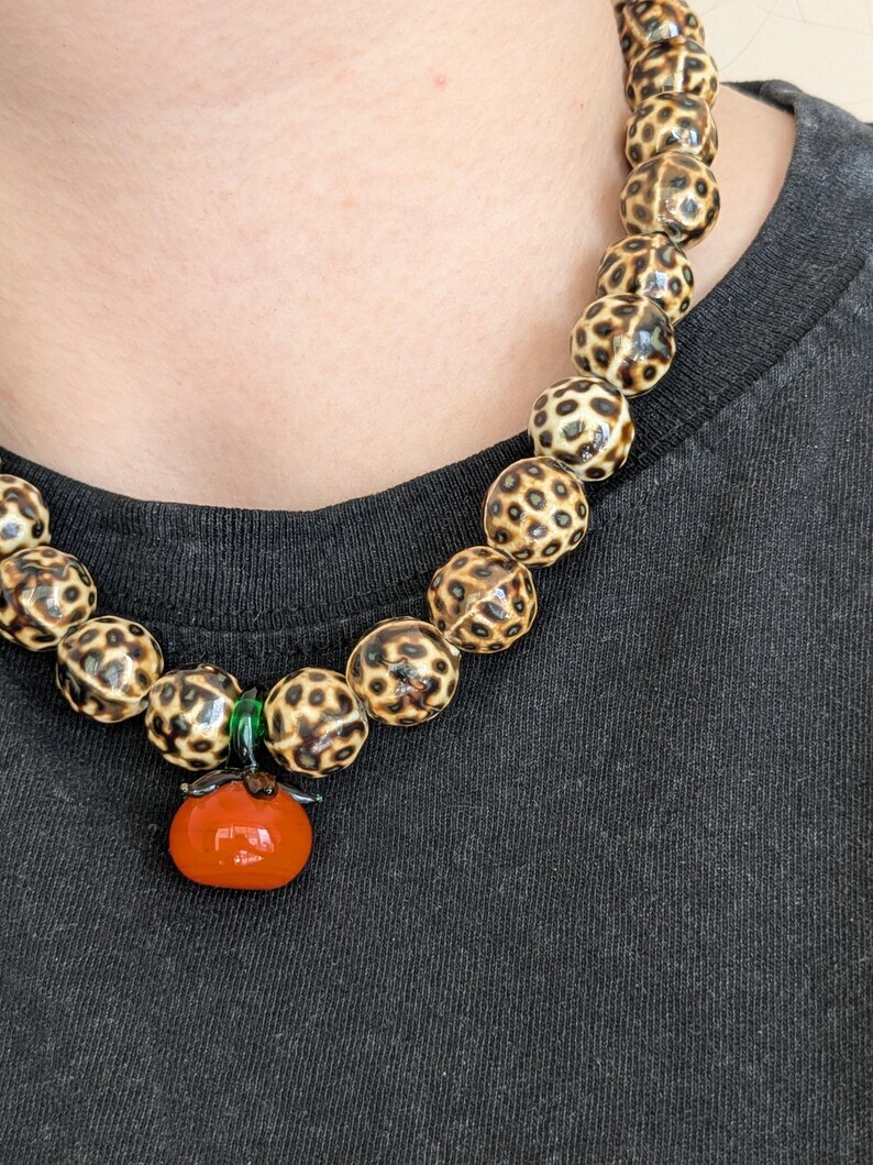 May include: A beaded necklace featuring large, round beads with a spotted pattern in shades of brown and cream. The necklace has a pendant of a small, orange tomato with green accents.