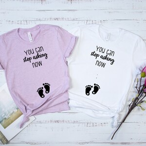 Puede incluir: Dos camisetas con el texto "You can stop asking now" y un par de huellas de pies de bebé. Una camiseta es blanca y la otra es de color morado claro.