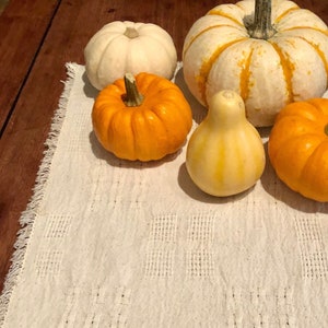 May include: Five gourds on a white crocheted table runner. The gourds are various shapes and sizes, including a white gourd with yellow stripes, a yellow gourd, and three orange gourds.