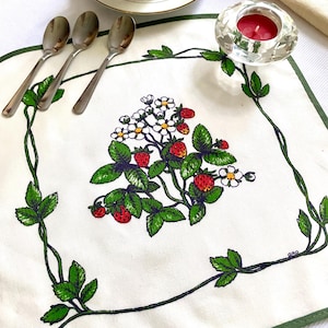 May include: A white placemat with a green border features a strawberry design with red berries, green leaves, and white flowers. Three silver spoons are on the left. A crystal candle holder with a red candle is on the right.