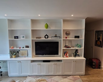 May include: A white built-in entertainment unit with a flat screen TV, shelves, and cabinets. The unit is in a living room with a desk and chair in the foreground.