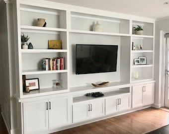 May include: White built-in entertainment unit with shelves, cabinets, and a flat screen TV. The unit has a wood-look floor and a white wall behind it.