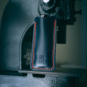 Könnte beinhalten: Ein dunkelblaues Lederetui mit roten Nähten und einem kleinen Logo mit drei Pfeilen. Das Etui hängt an einer Metallpresse.