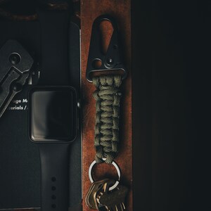 May include: A black metal carabiner with a green paracord wrapped around it. The carabiner is attached to a silver key ring with a set of keys. The carabiner and keys are sitting on a brown leather surface.