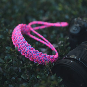 May include: A pink and blue braided camera strap with a black camera lens in the background.