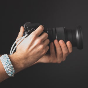 May include: A white and blue braided wrist strap for a camera. The strap is made of a thick, durable rope and has a blue and white pattern.