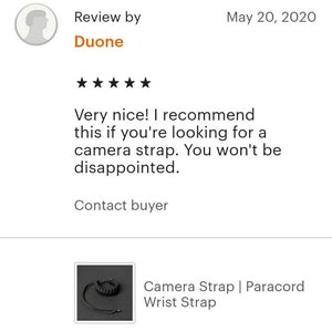 May include: A black camera strap made of paracord. The strap is adjustable and has a buckle closure. The strap is shown wrapped around a wrist.