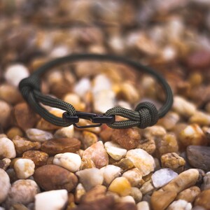 Pode incluir: Um bracelete de paracord verde com um fecho de metal preto. O bracelete est&aacute; sobre um leito de pequenas pedras marrons.