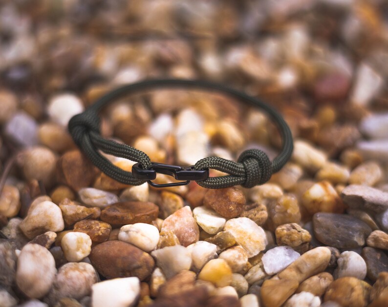 Pode incluir: Um bracelete de paracord verde com um fecho de metal preto. O bracelete est&aacute; sobre um leito de pequenas pedras marrons.