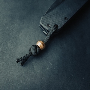 May include: A black folding knife with a black paracord lanyard and a wooden bead. The knife is lying on a dark gray surface.