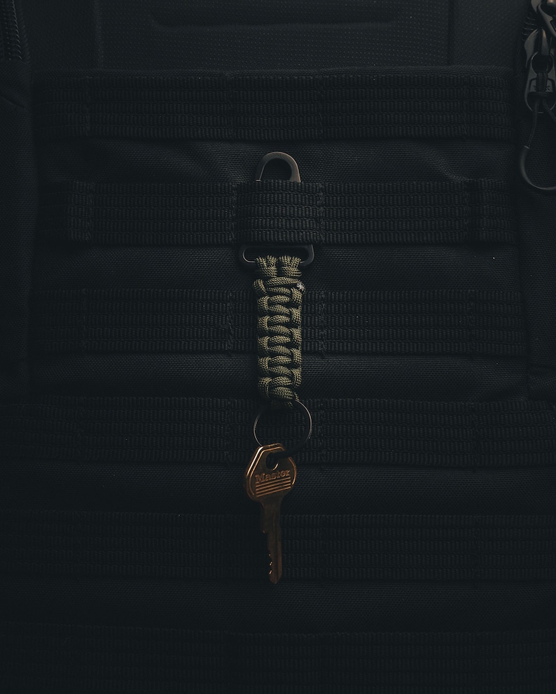 May include: A green paracord keychain with a gold key attached to a black tactical backpack.